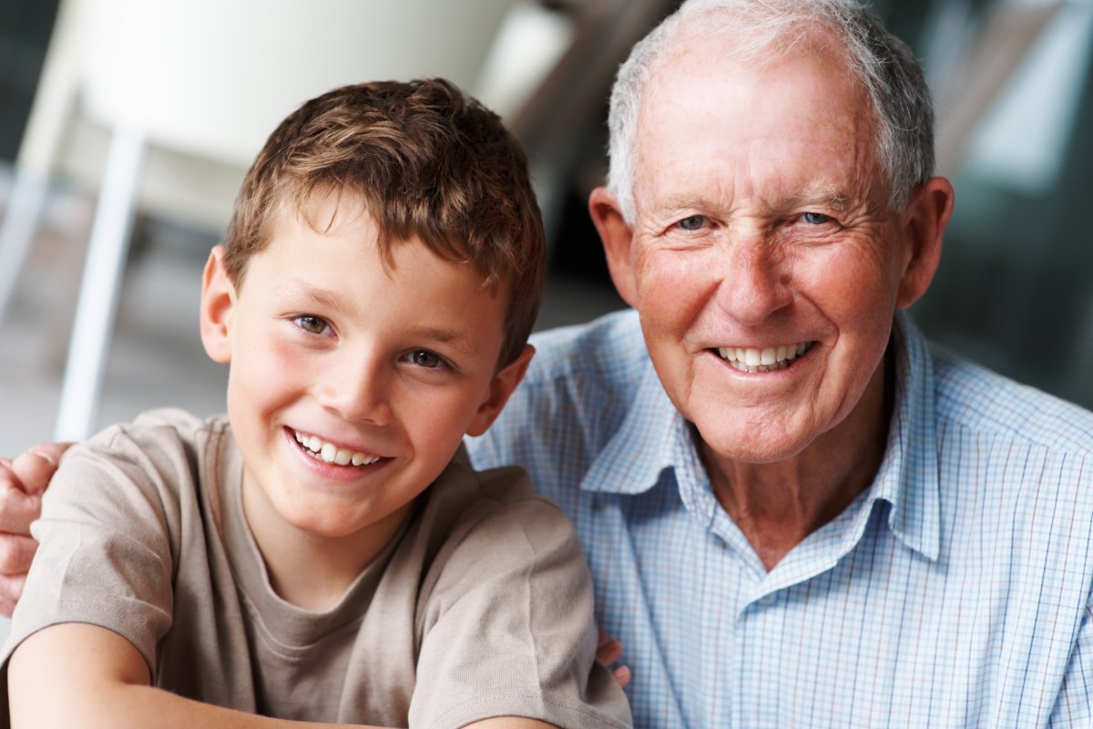 Close-up of an elderly man wearing a blue button-down with his arm over a young boy's shoulder wearing a brown t-shirt
