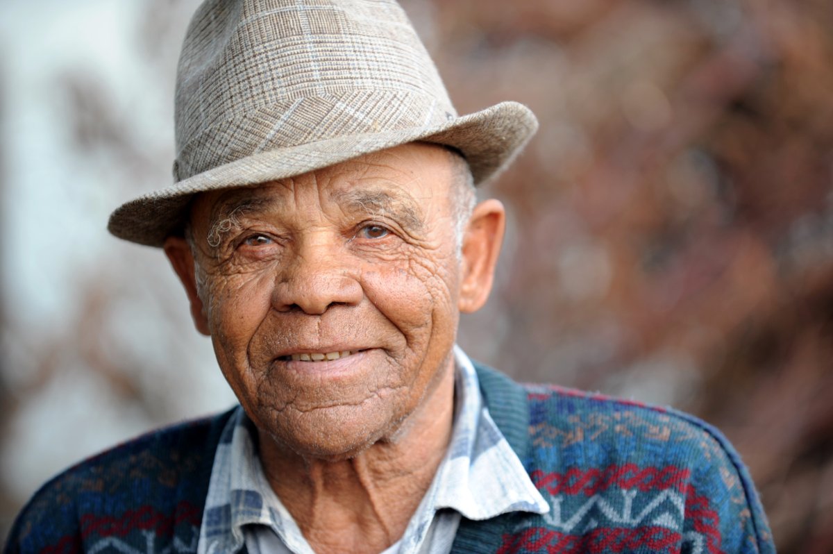 A close-up of an elderly man wearing a plaid fedora hat and a patterned sweater.