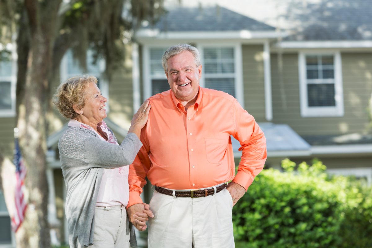 A close-up of a couple holding hands in front of a house with an American flag. The man wearing an orange button-down and khakis and the woman is wearing a pink polo with a gray cardigan and dress pants.