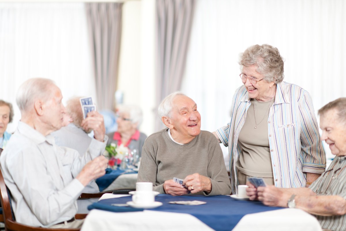 A group of seniors is sitting around a table in a dining room, playing cards. Gray curtains, tan walls, and blue table cloth on the table