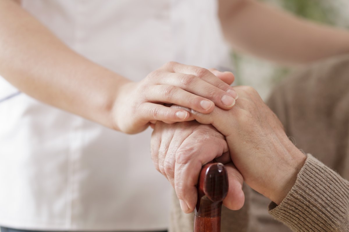 A close-up of a senior holding on to a cane with a caregiver having their hand over the senior's hand. The caregiver is wearing white scrubs.