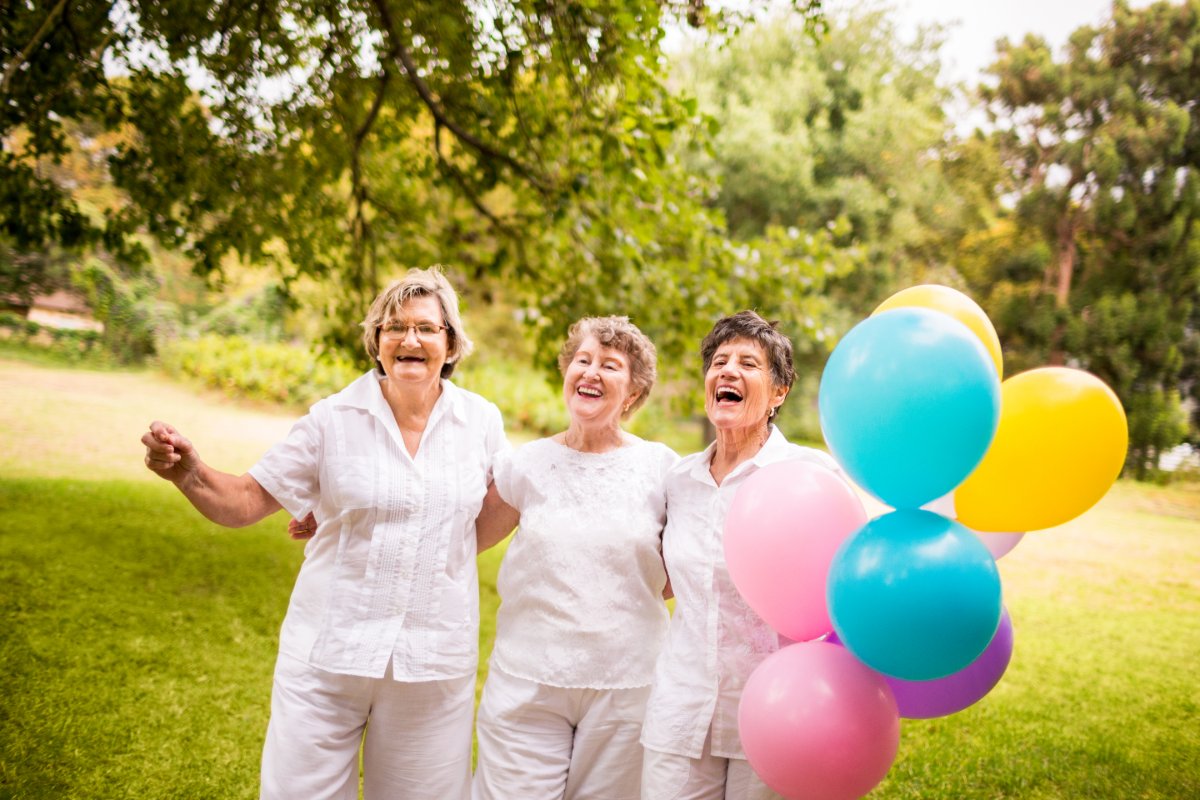 A group of three women outside wearing white. One woman is holding bright-colored balloons.