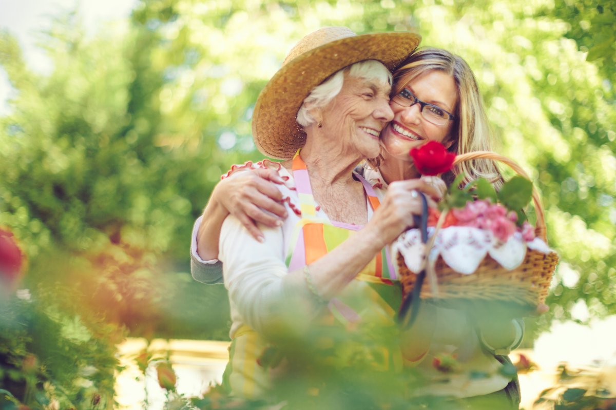 2 women are outside picking flowers in the garden. An elderly woman is wearing a straw hat and holding a rose and the other woman has her hand across the elderly woman's shoulder and holding a basket of flowers.