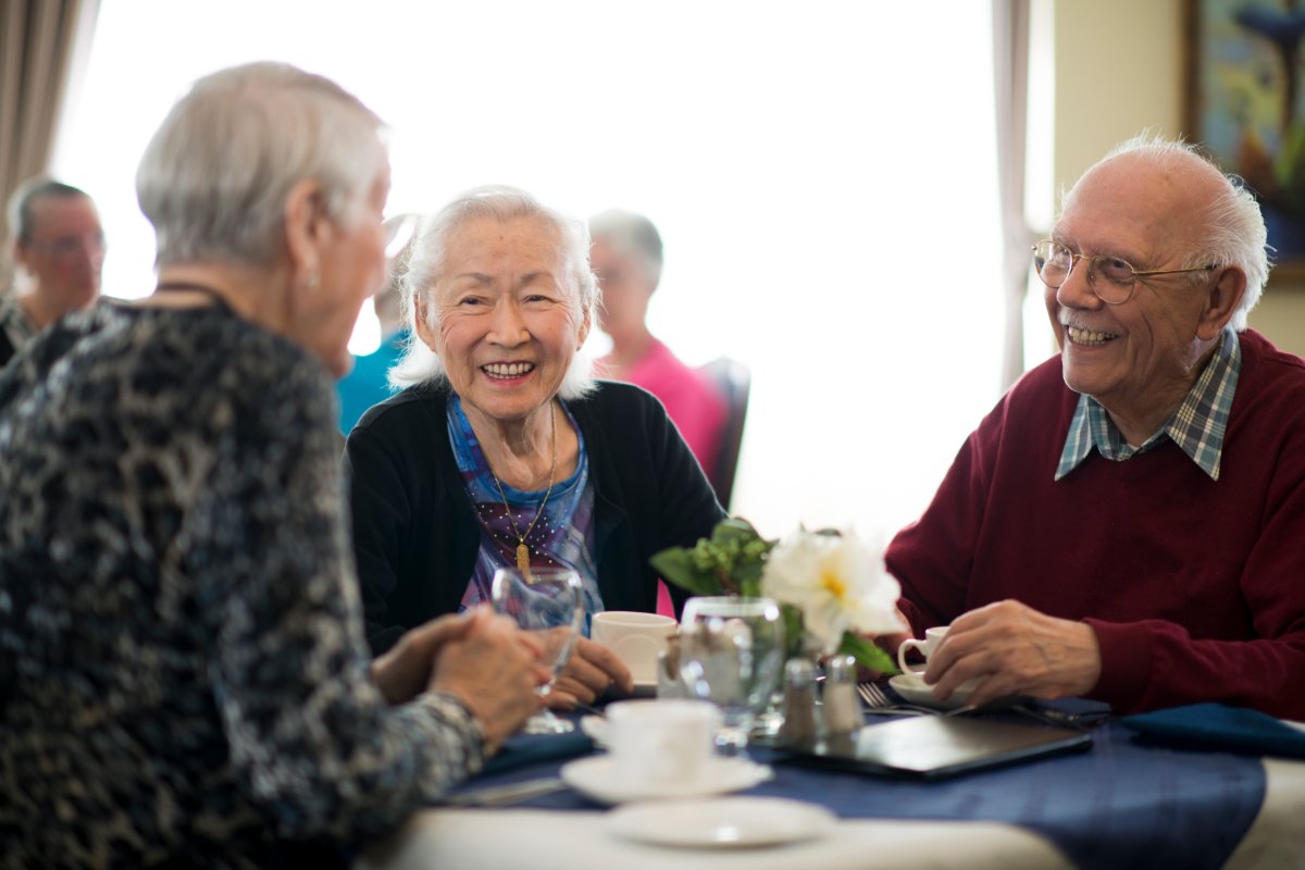 A group of senior citizens are sitting around a dining room table. The table is white with a blue tablecloth. The table has coffee cups, salt and pepper, water glasses, and silverware.