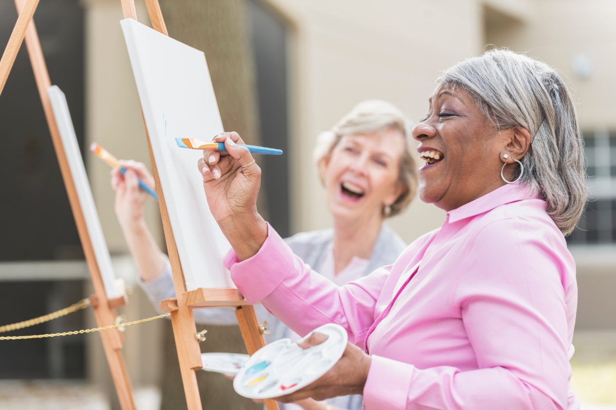 Two elderly women outside painting canvas. Both hold a paint palette and hold up a brush to the canvas.