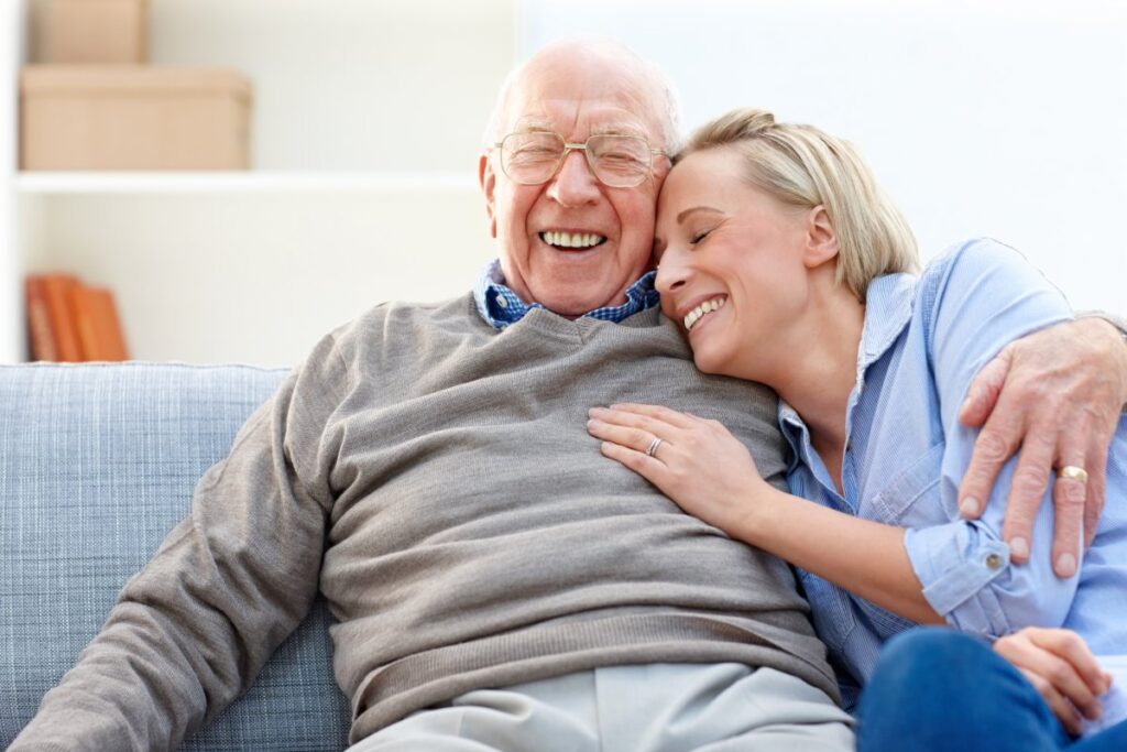 A senior man and his daughter are laughing and hugging on the couch