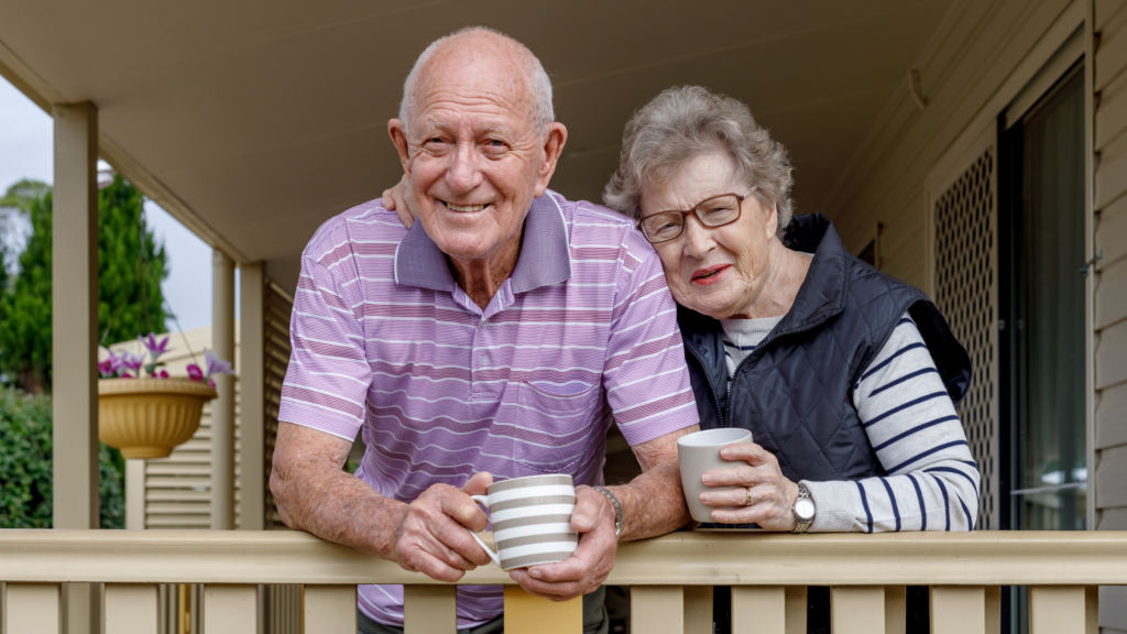 A senior man and senior woman standing together on a porch leaning against the railing while holding coffee mugs