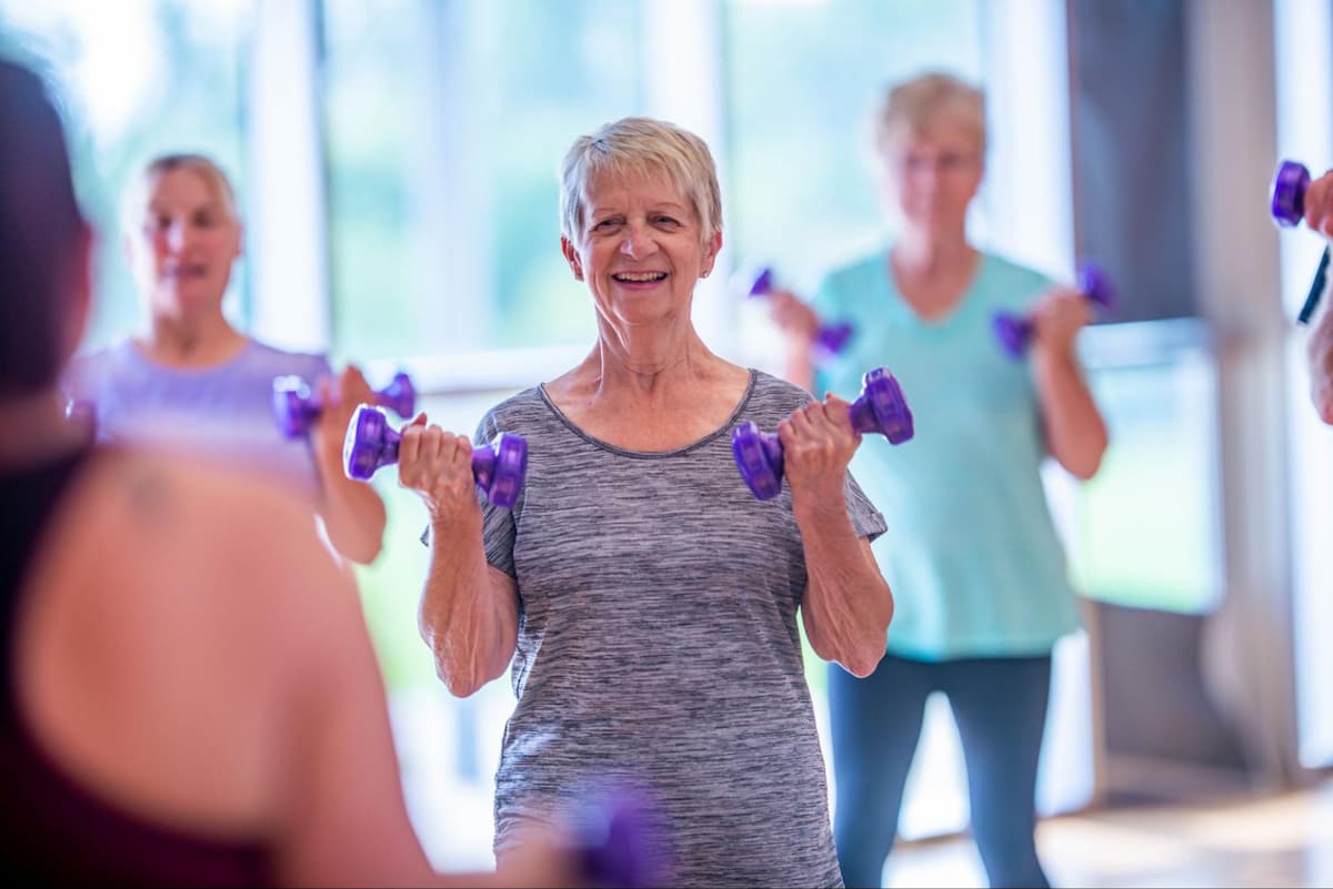Three senior women wearing workout clothes and exercising with small purple weights while they look at an instructor